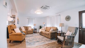 Un salon avec un fauteuil en cuir marron, un canapé beige et une chaise en bois. On y trouve un tapis à motifs sur le sol, une horloge au mur et une porte avec une fenêtre.