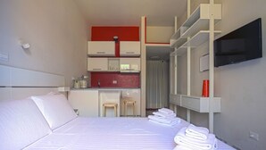 Quarto de hotel com cama, cozinha compacta, televisão e um pequeno espaço para sentar.