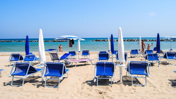 Ein Strand mit Reihen aus blauen Liegestühlen und weißen Sonnenschirmen.