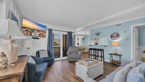 Een woonkamer met een houten vloer, een blauwe bank, een witte salontafel en een televisie waarop een strand te zien is.
