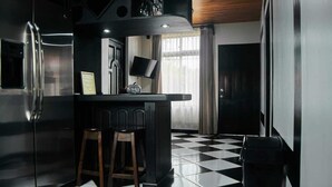 Uma cozinha moderna com piso quadriculado em preto e branco, uma geladeira de aço inoxidável, um balcão com banquetas e uma TV de tela plana presa na parede.