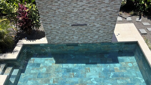 Uma piscina retangular com borda em mosaico de pedra e parede de pedra ao fundo.