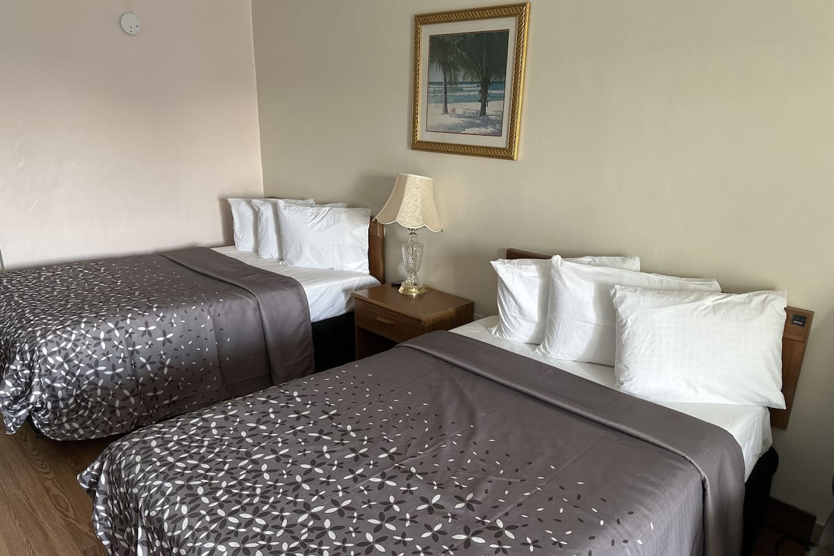 Une chambre d’hôtel avec deux lits simples, chacun recouvert d’une couvre-lit à motifs floraux gris et agrémenté d’oreillers blancs. Une table de chevet avec une lampe se trouve entre les deux lits.