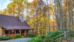 Una cabaña rústica con chimenea de piedra, rodeada por un bosque con follaje otoñal.