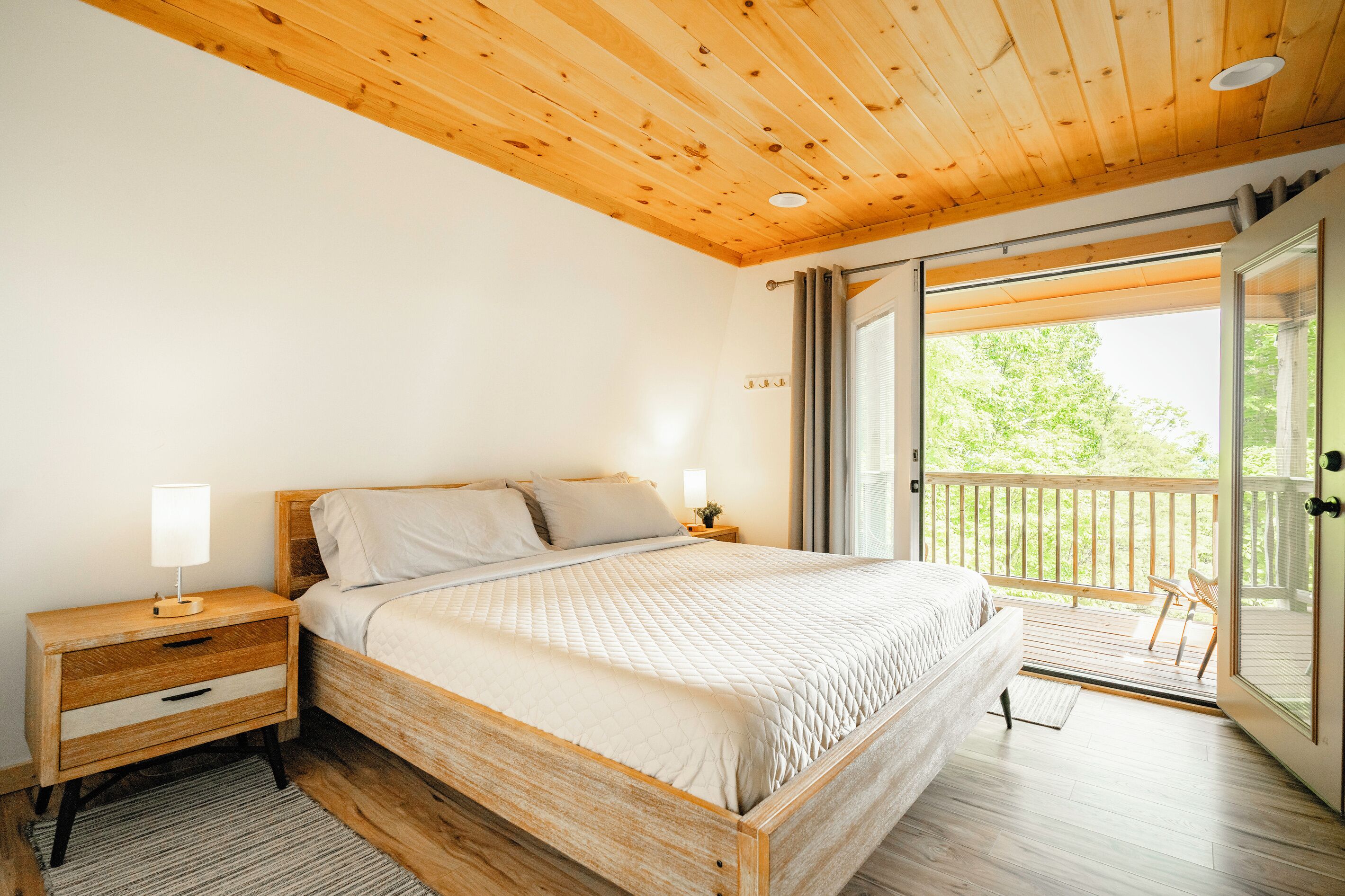 Une chambre avec un plafond en bois, un lit recouvert d’une courtepointe matelassée, une table de chevet avec une lampe et une porte-fenêtre coulissante donnant sur une terrasse extérieure.