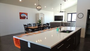 A modern kitchen with a central island, dark cabinetry, and a dining area with chairs and a table. - Red Rock Oasis and Private Pool Getaway! (Moab)