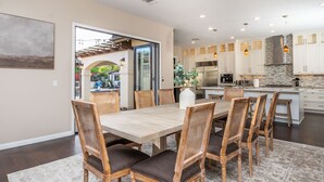A spacious dining area with a large wooden table and wicker chairs, a kitchen with white cabinets and a stone backsplash, and a view through a glass door to an outdoor patio. - Hidden Gem of SoCal: Game Room, Pool, Spa, & Resort Backyard!  (Vista)