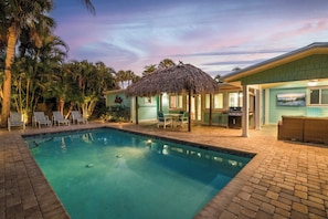 A swimming pool with lounge chairs, a thatched roof structure, and a building with a view of the water.