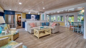 Amplia sala de estar con sofá, mesa de centro y mesa de comedor. Se ve una cocina con refrigerador y lavadora al fondo.
