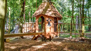 A wooden birdhouse structure in a forested area with a rope bridge and a wooden play structure nearby.