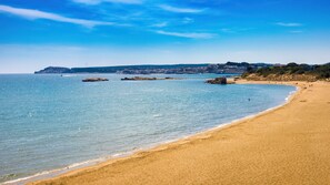 Une plage à l’eau d’un bleu limpide, avec un rivage sablonneux et une ville qui se devine au loin.