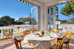 Un espace extérieur aménagé pour les repas, avec une table dressée pour quatre personnes, entourée de chaises en osier, d’un parasol vert et offrant une vue sur le paysage environnant.
