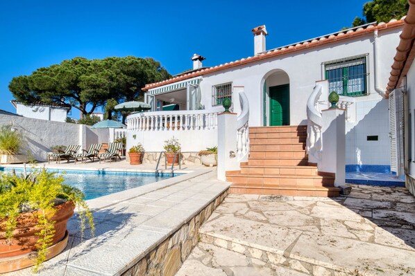 A white villa with a swimming pool, outdoor seating, and a staircase leading to a green door.