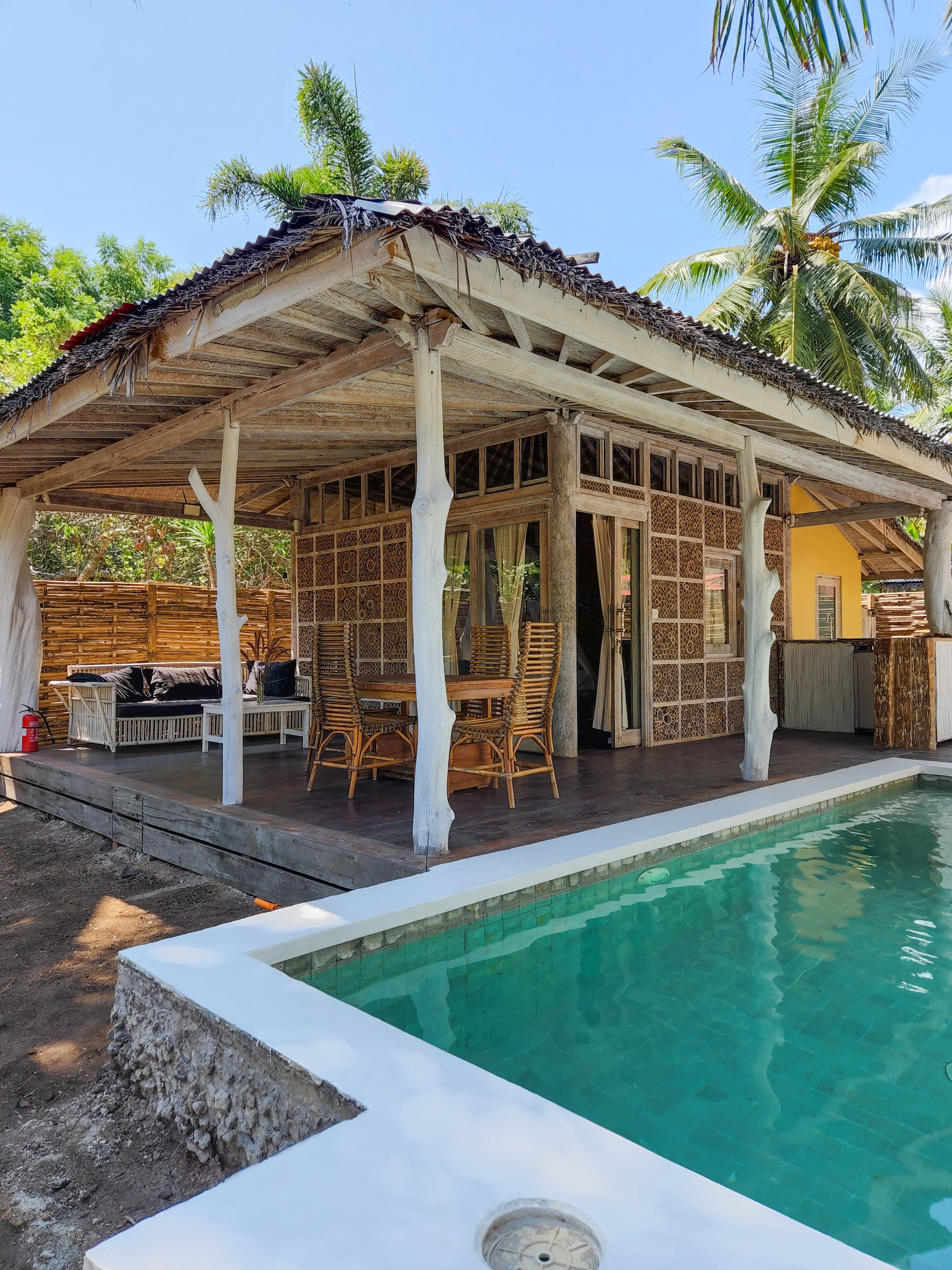 A covered outdoor area with a pool, wooden furniture, and a thatched roof.
