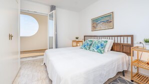 A bedroom with a bed, a nightstand, a wall-mounted shelf, a painting, and a view through a door. - Pleasant apartment with sea view (Empuriabrava)