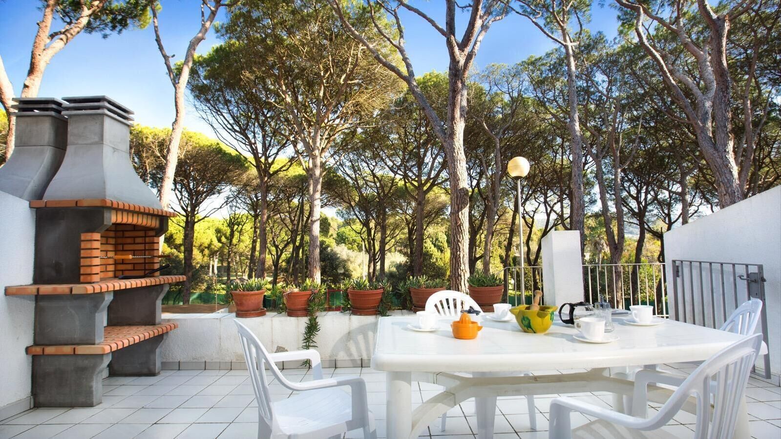 Eine Terrasse im Freien mit einem Backstein-Grill, weißen Stühlen, einem Tisch und Topfpflanzen.