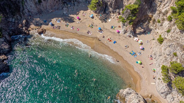 Vista aérea de una playa con aguas turquesas cristalinas, acantilados rocosos y numerosos paraguas de playa.