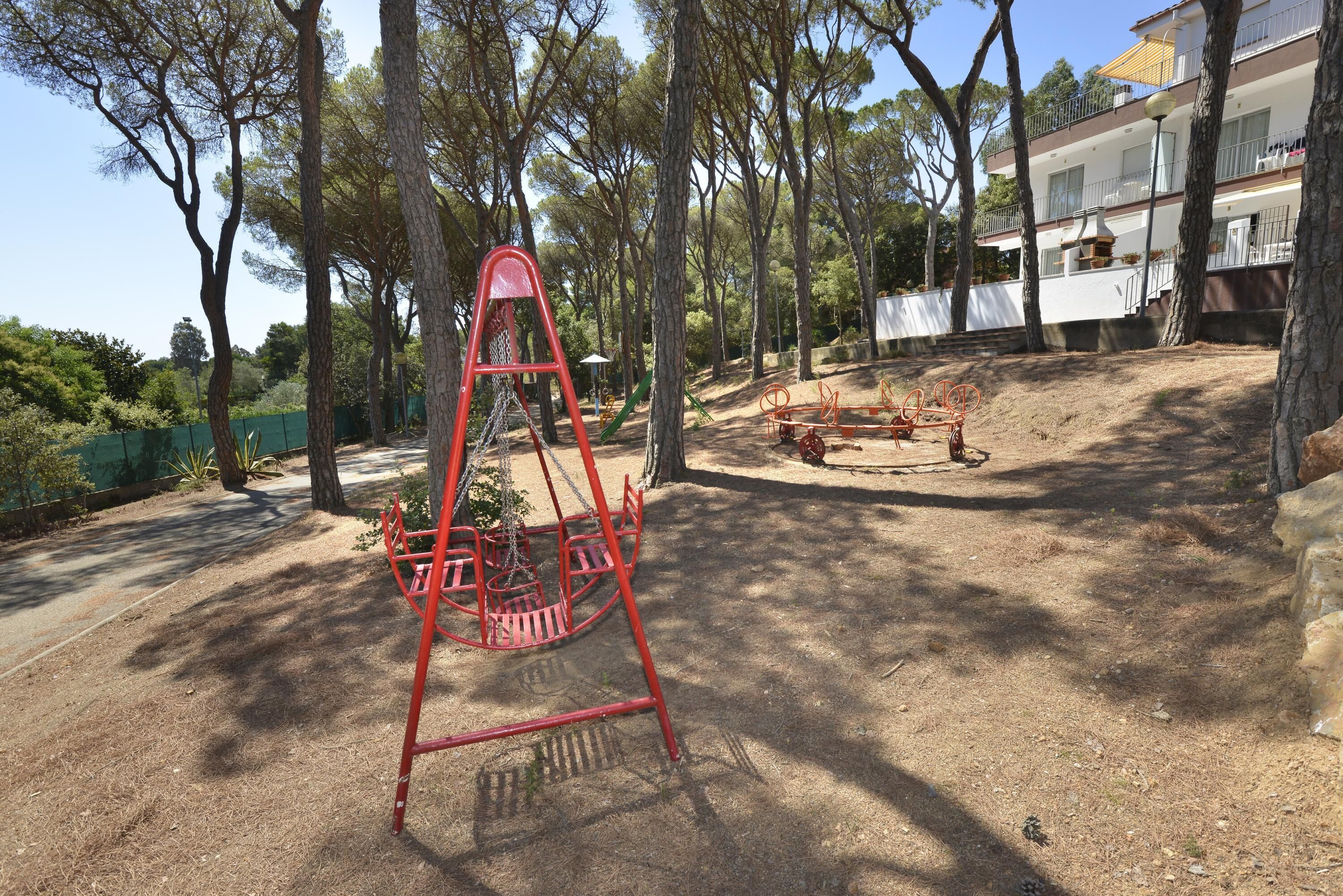 Un’area giochi con altalene e uno scivolo, circondata da alberi e un edificio sullo sfondo.