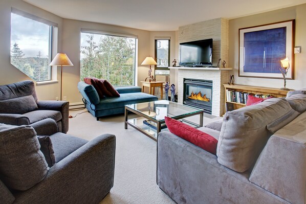 A modern living room with a fireplace, comfortable seating, and large windows offering a view of trees. - Ski in/Out 2 Bed/2bath w Pool/HotTub & Great Light (Whistler)