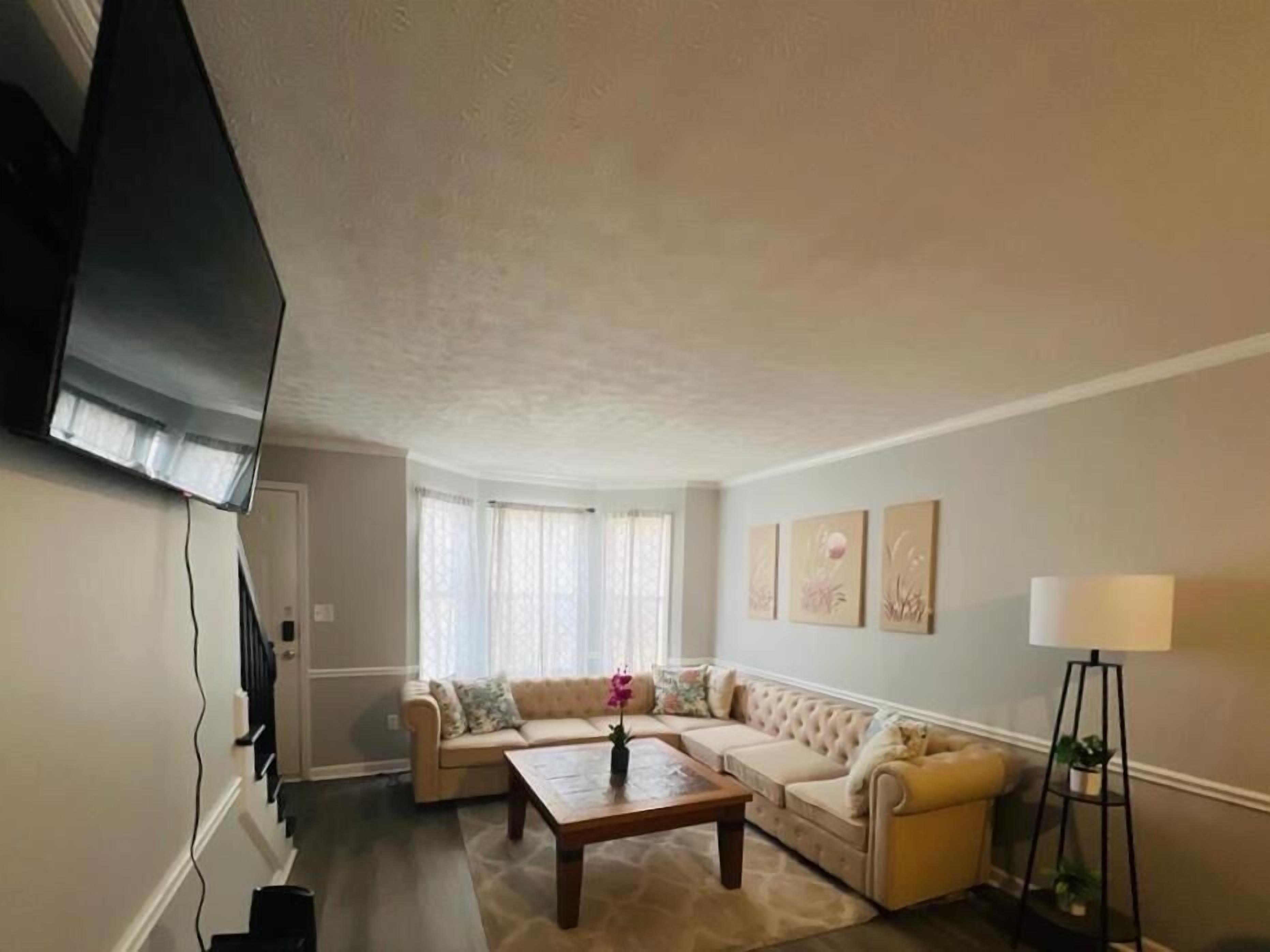 A living room with a large flat-screen TV mounted on the wall, a beige sectional sofa, a wooden coffee table, and a floor lamp.