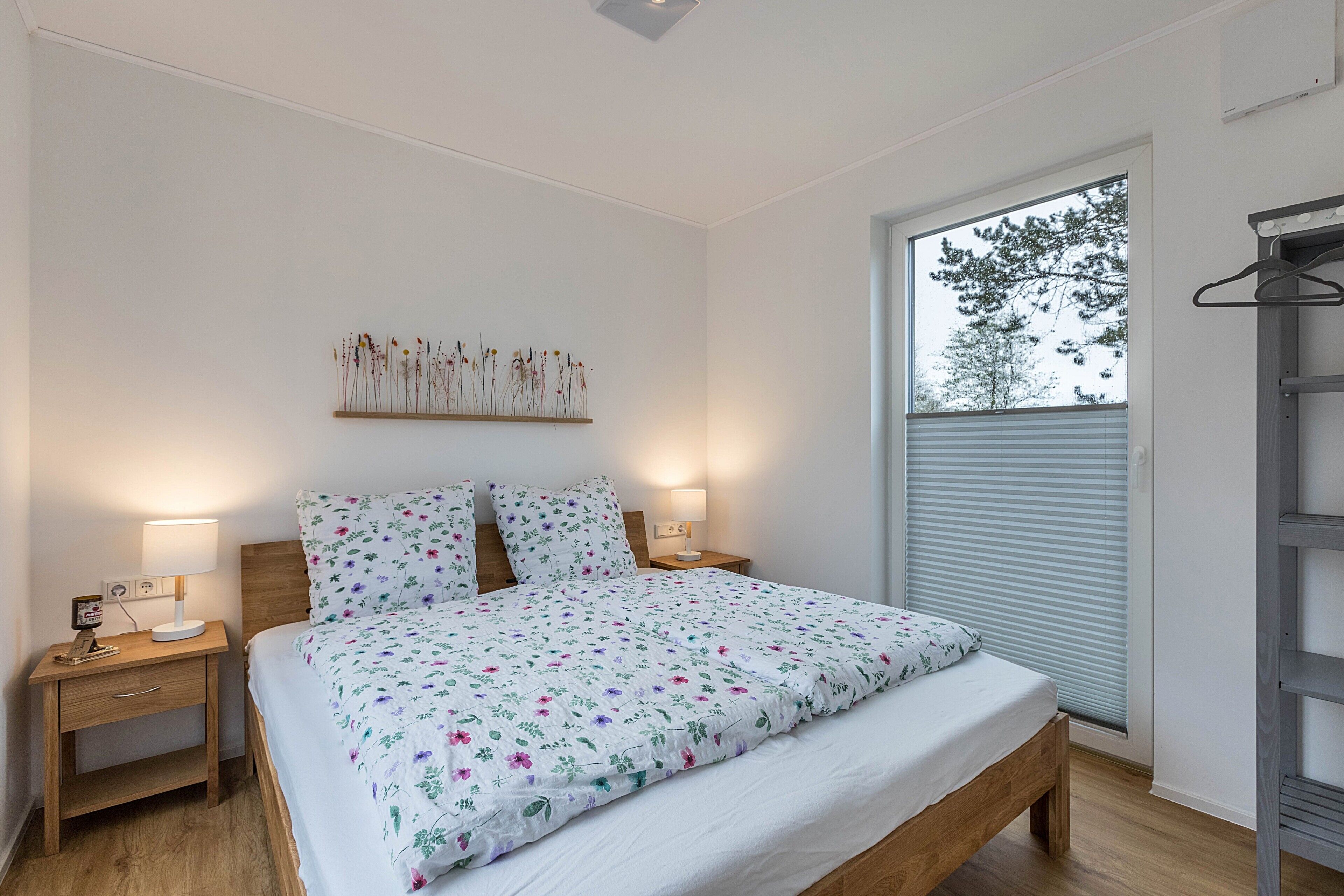A bedroom with a bed, bedside tables, a wall-mounted shelf with dried flowers, a window with blinds, and a wooden ladder.