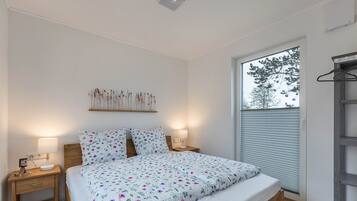 A bedroom with a bed, bedside tables, a wall-mounted shelf with dried flowers, a window with blinds, and a wooden ladder.