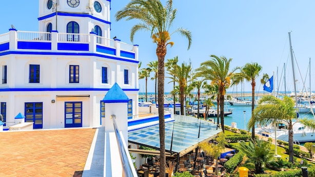 Un immeuble à plusieurs étages aux couleurs bleu et blanc, une terrasse sur le toit avec des espaces de détente en plein air, et une marina avec des bateaux en arrière-plan.