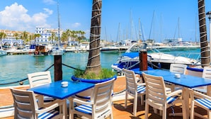 Un port de plaisance avec des bateaux, des palmiers et un restaurant aux tables bleues et aux chaises blanches.