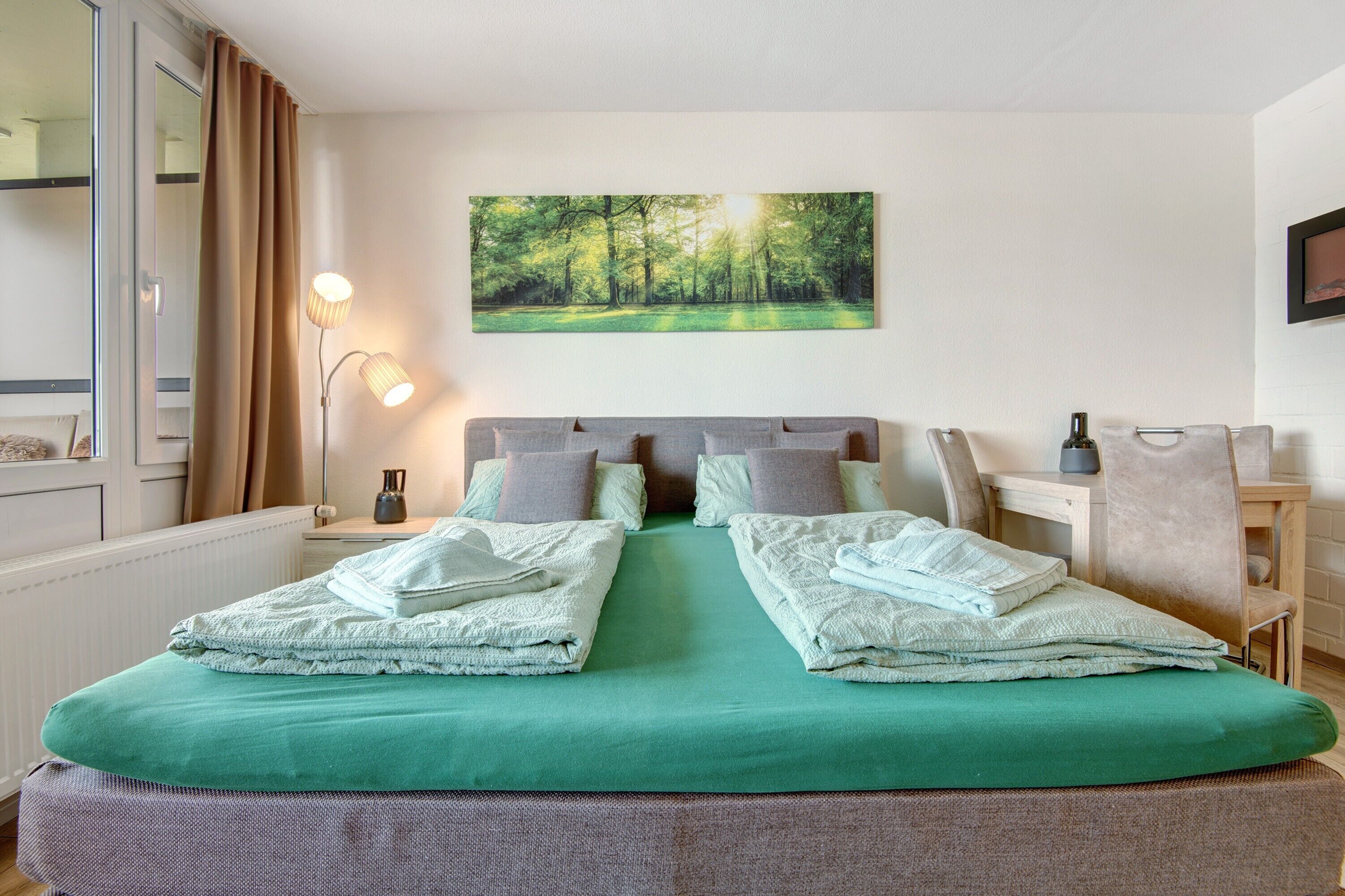 Un lit double avec une literie verte, une tête de lit, deux lampes de chevet, un tableau représentant une forêt et une chaise avec une serviette.