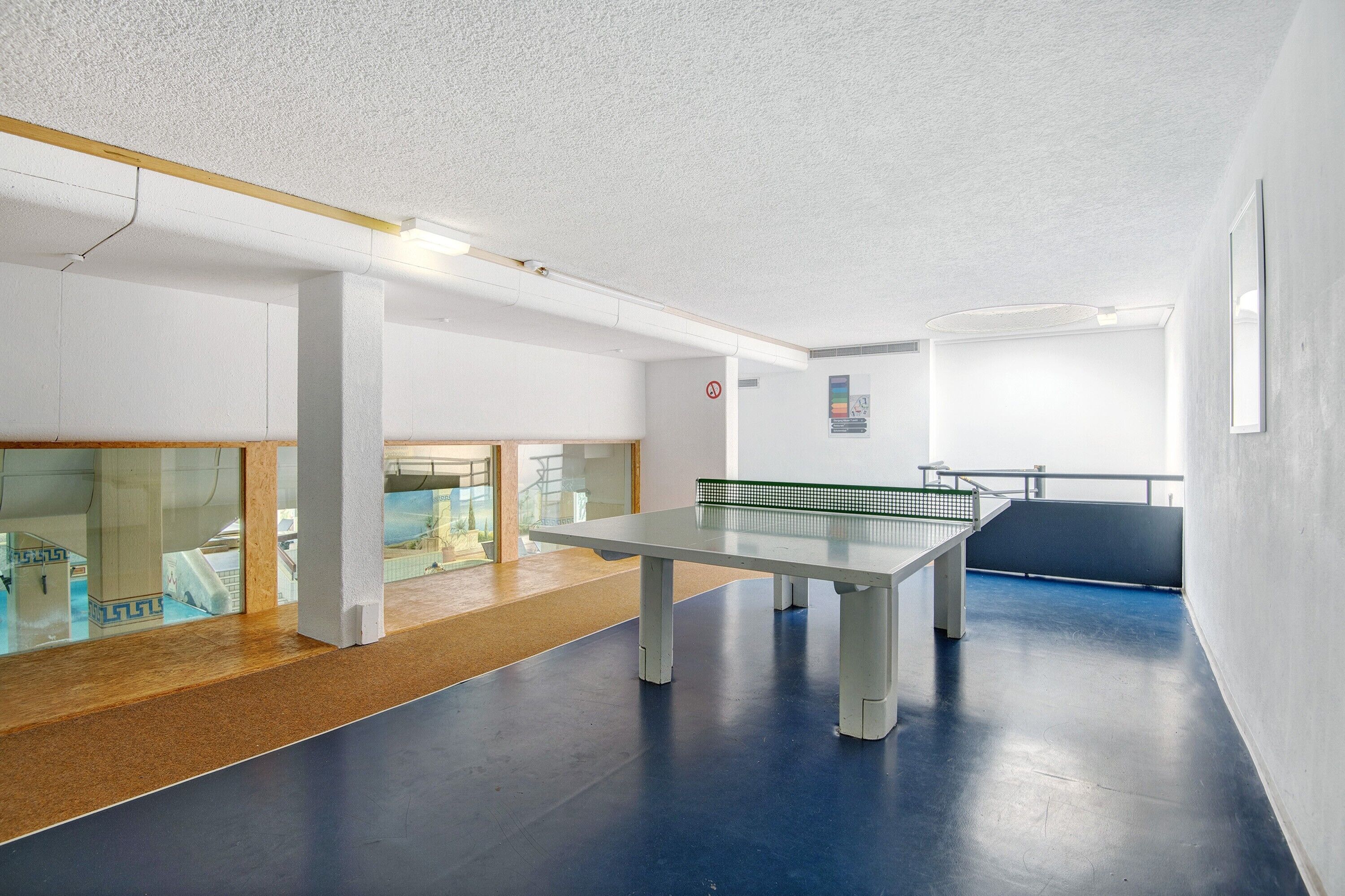 A ping pong table in a room with white walls, a blue floor, and a glass partition.