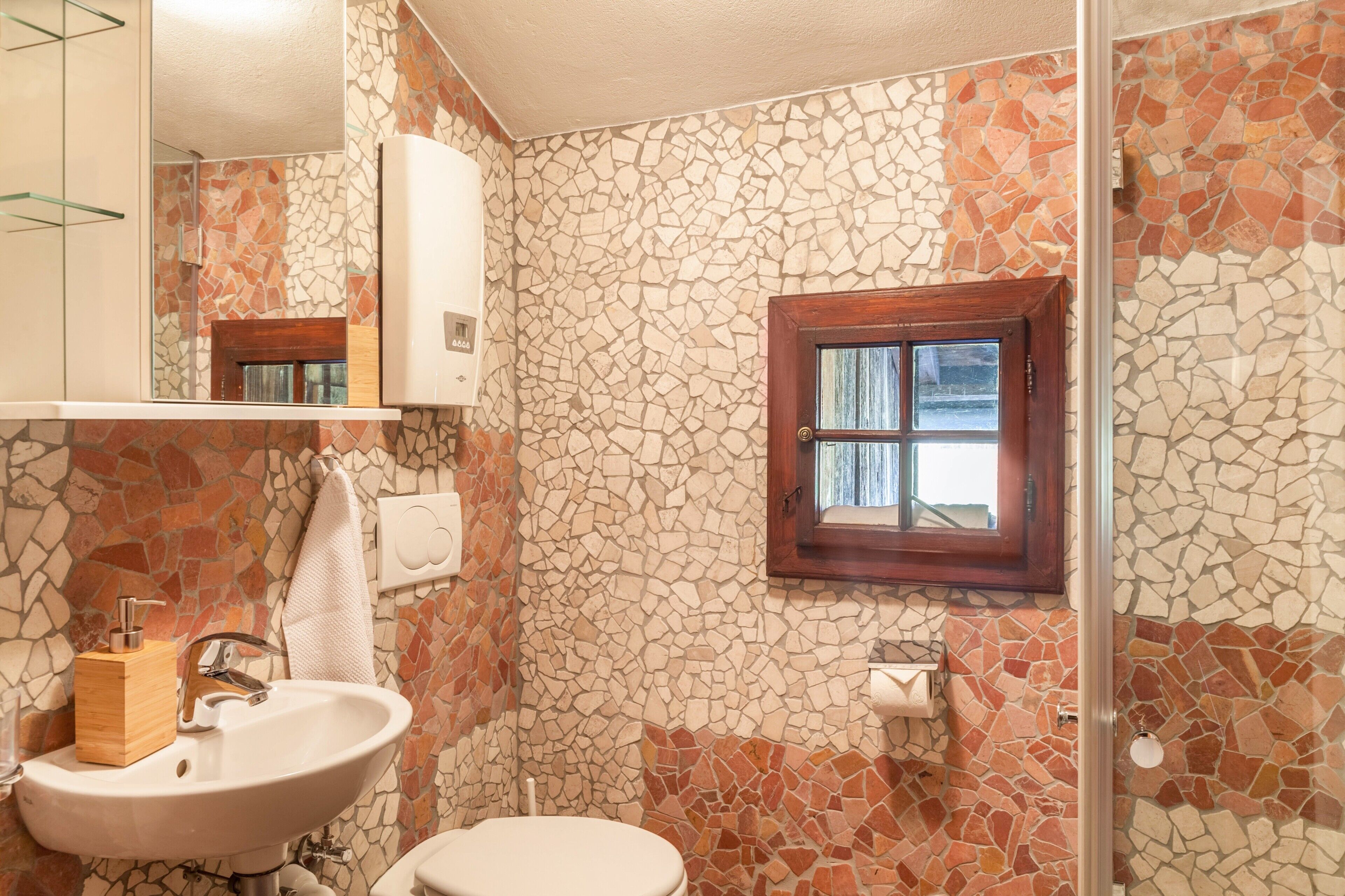A bathroom with a stone mosaic wall, a wooden window, a white sink, and a toilet.