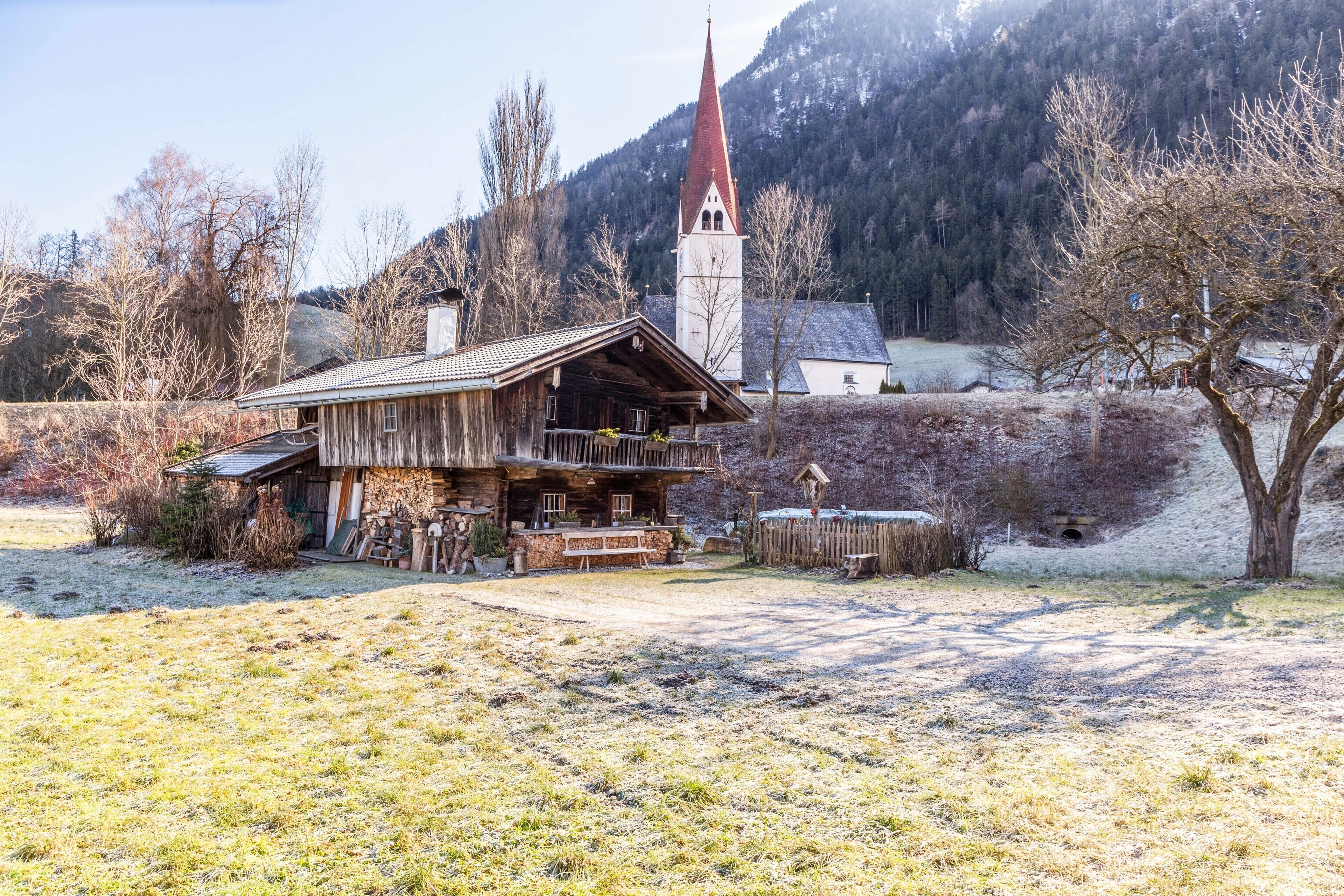 Ein rustikales Holzgebäude mit Balkon, eine Kirche mit rotem Kirchturm und eine gebirgige Kulisse.