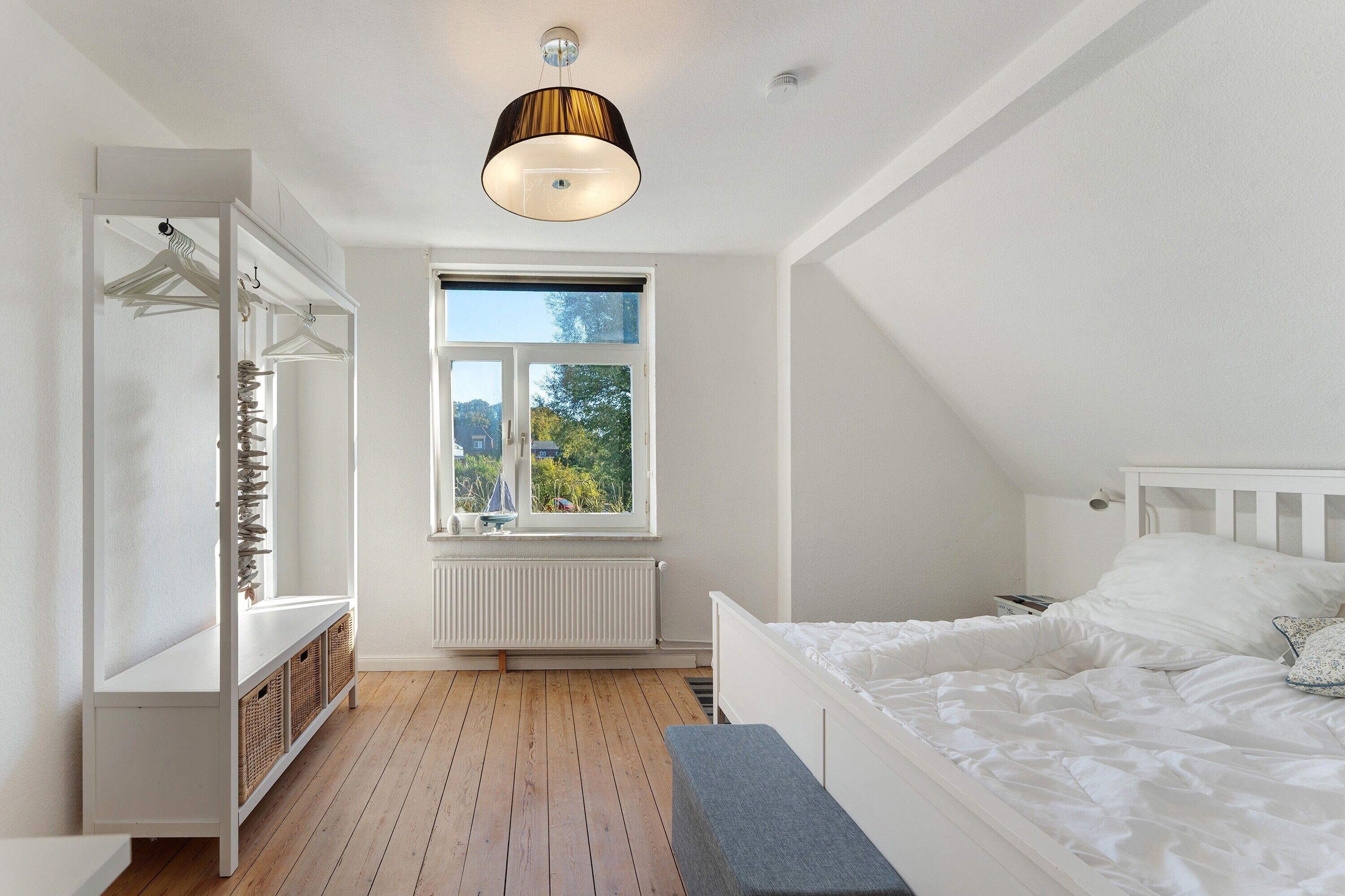 Un dormitorio con una cama blanca, una ventana con vistas, un armario blanco y una lámpara colgante.