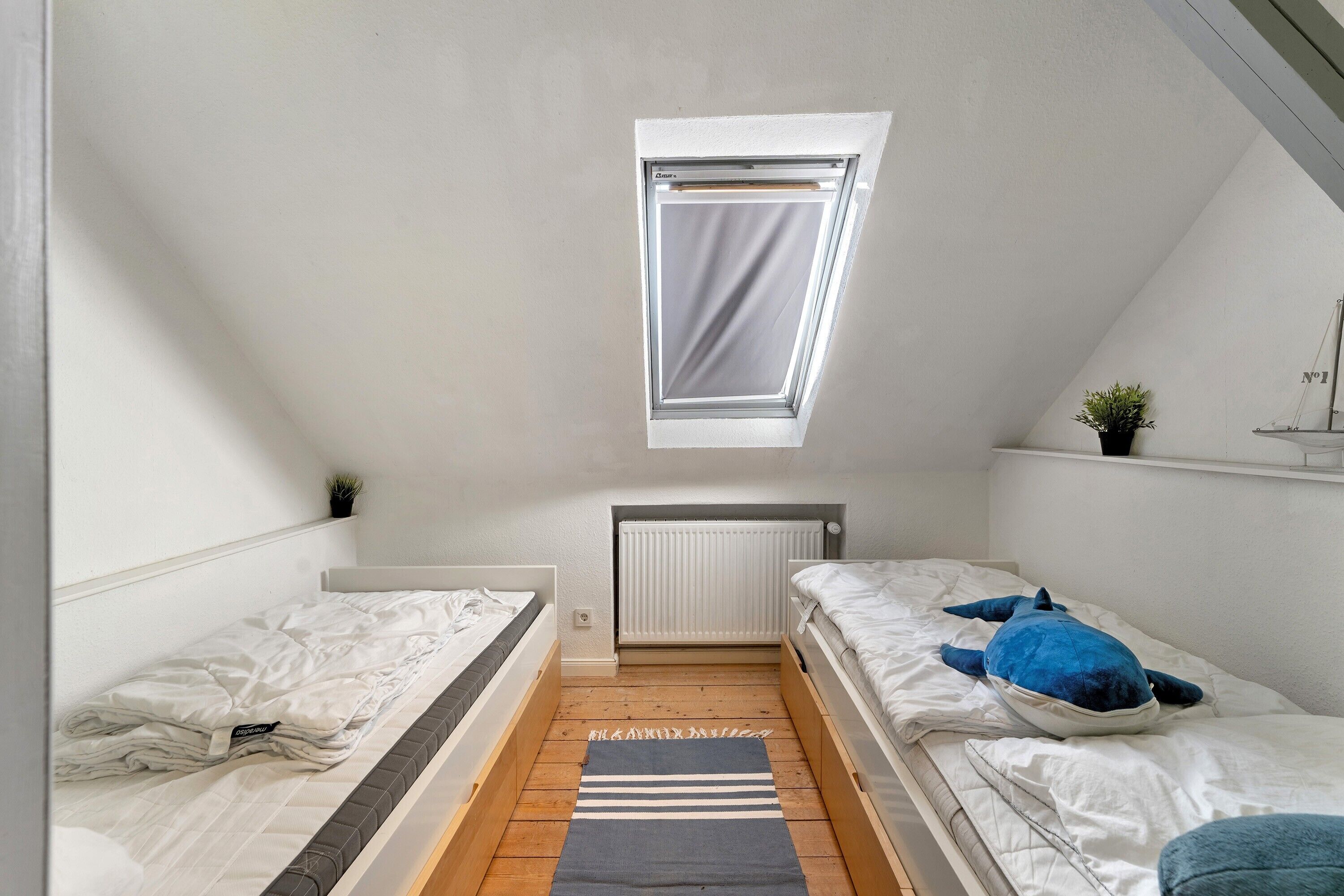 Un dormitorio ático pequeño y triangular con dos camas, un tragaluz, un radiador y una planta pequeña.