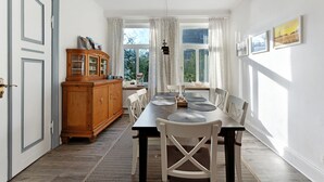 A dining room with a wooden cabinet, a dining table with chairs, and a window with curtains. - Vacation Home "Insel Hus" with Private Terrace, Garden & Wi-Fi (Fehmarn)