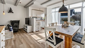 A modern kitchen with white cabinets, a dining area with a wooden table and chairs, and a large window providing a view of the outdoors. - Vacation Home "Deich Hus" with Private Terrace, Garden & Wi-Fi (Büsumer Deichhausen)
