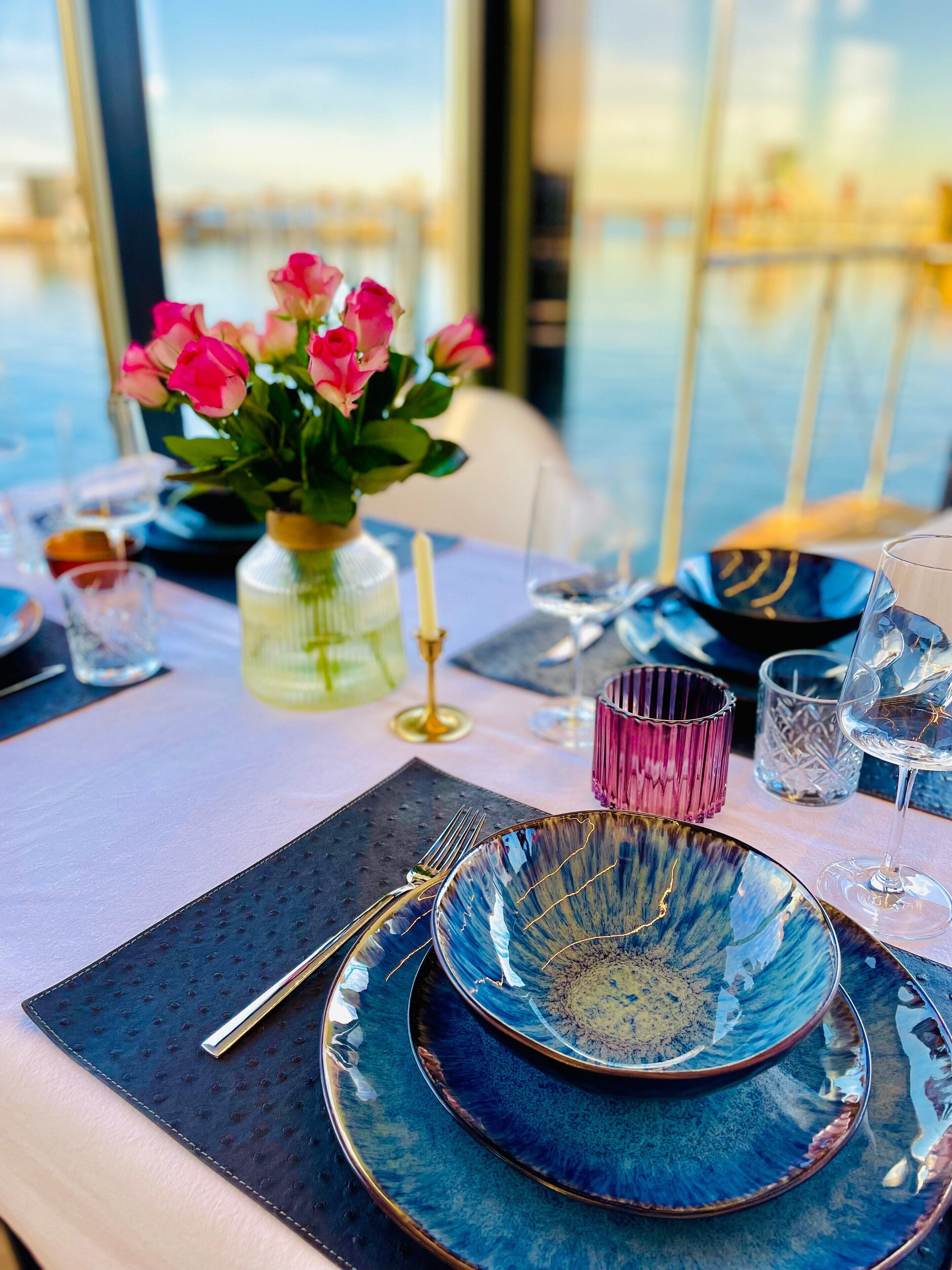 A dining table with blue plates, a vase with pink flowers, and a view of the water outside.