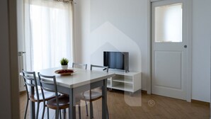 Un comedor moderno con una mesa blanca, cuatro sillas, una pequeña planta en maceta y un televisor sobre un mueble blanco.