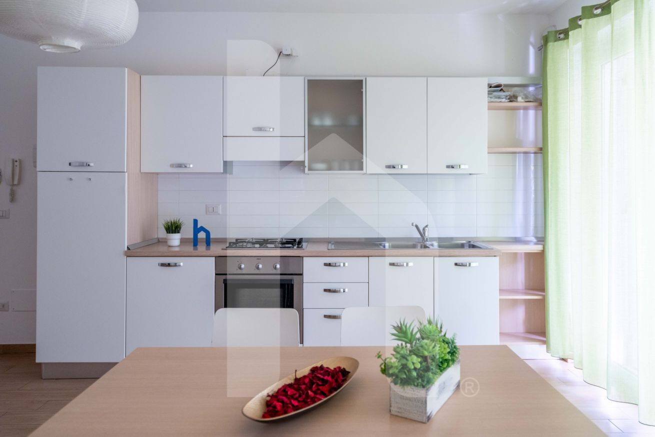 Una cucina moderna con mobili bianchi, un'isola centrale e un tavolo da pranzo con una pianta e una ciotola di fiori rossi.