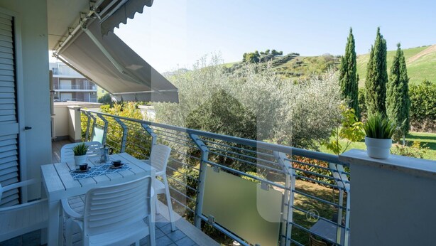 Un balcón con mesa y sillas, con vista a un paisaje montañoso con árboles y un edificio al fondo.
