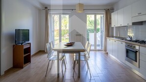 Une cuisine moderne avec des meubles blancs, une table à manger avec des chaises, un téléviseur posé sur un meuble et une grande fenêtre avec des rideaux.