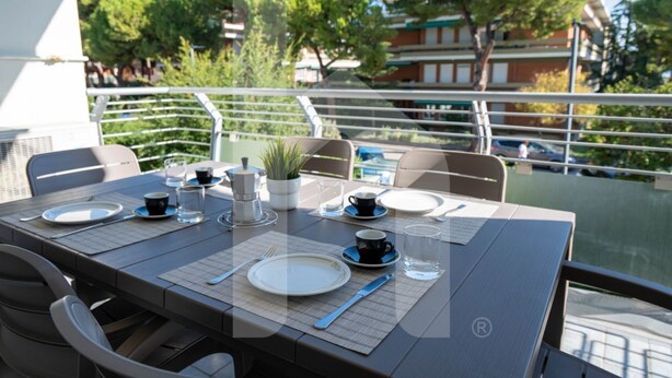 A dining table set with plates, cutlery, and glasses, overlooking a body of water with a railing and greenery in the background. - Karismatico - Three-room apartment on the first floor a few steps from the sea (Pineto)