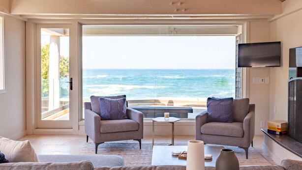 A living room with two armchairs, a coffee table, and a TV. It features a large glass door leading to a balcony with a view of the ocean.