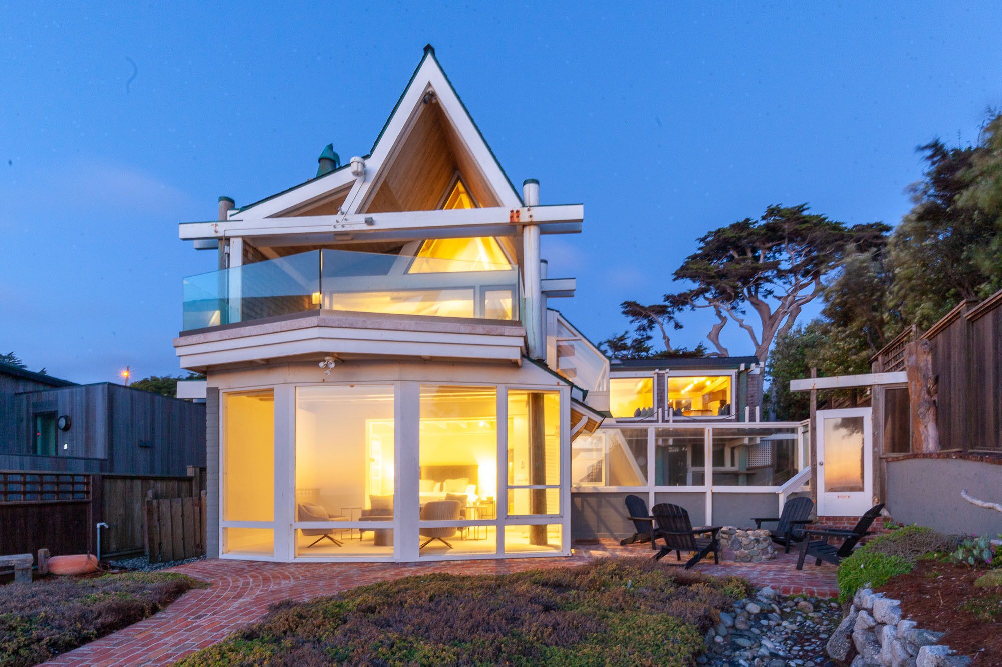 Une maison moderne à deux étages, dotée d’une façade en verre, d’un espace extérieur aménagé et d’un intérieur visible, avec une cheminée.