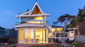 A modern two-story house with a glass facade, outdoor seating area, and a visible interior with a fireplace.