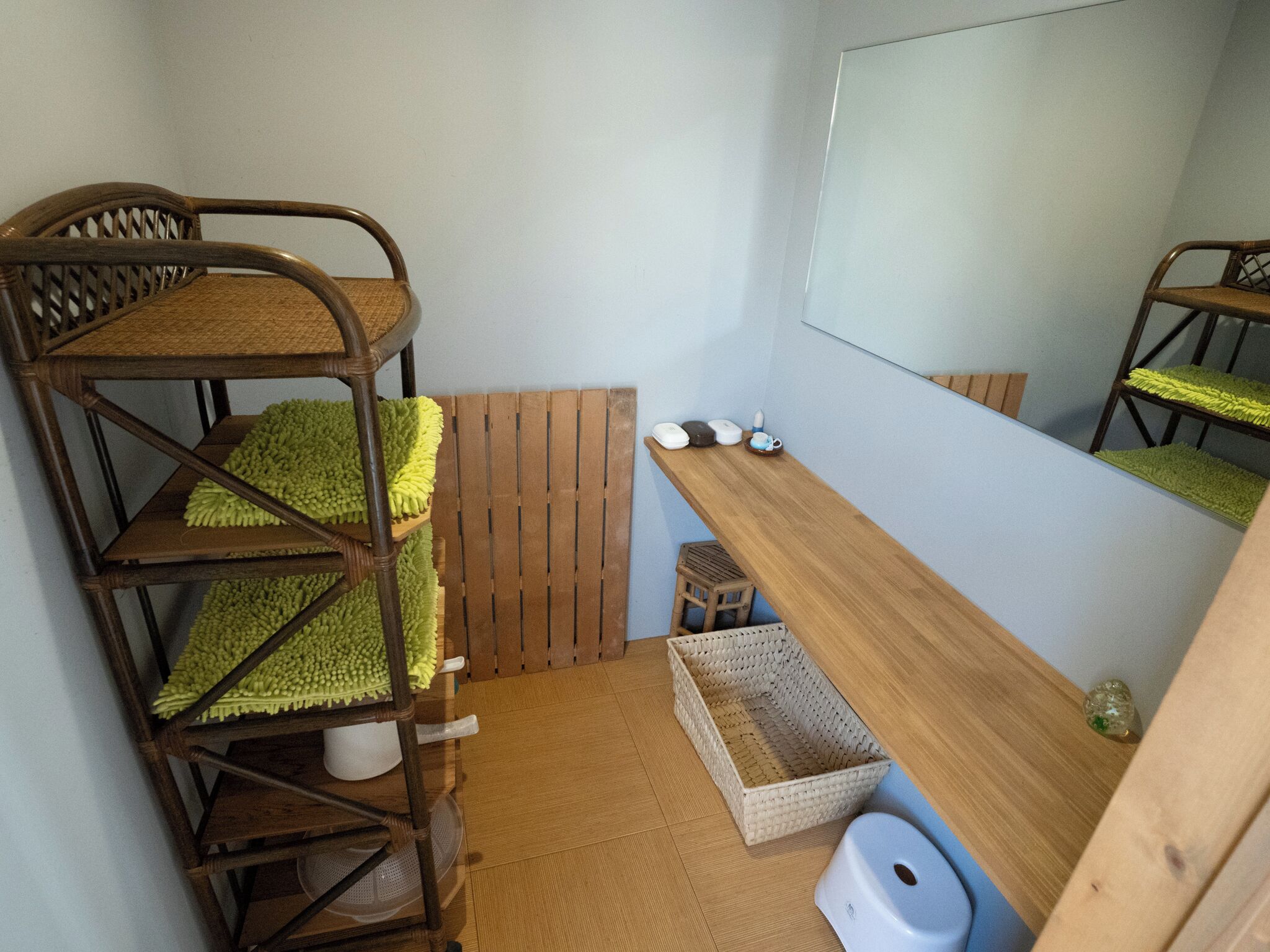 Una cama individual con estructura de madera y cojín de asiento tejido, un taburete pequeño de madera, una cesta tejida y un inodoro azul.