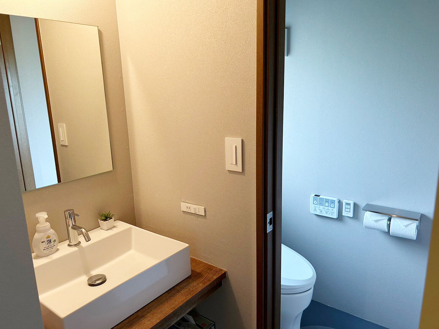 Un baño con lavamanos blanco, espejo y retrete. Hay dos interruptores de luz en la pared y un dispensador de papel higiénico.