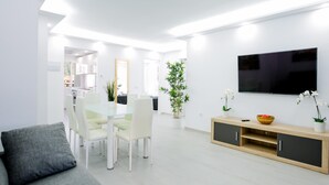Een moderne woonkamer met een grijze bank, een eethoek met een glazen tafel en stoelen, een aan de muur bevestigde televisie en een houten kast met decoratieve objecten.