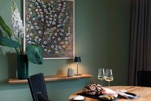 A dining area with a wooden table, a loaf of bread, olives, and a small bowl of cheese. There is a vase with flowers and a lamp on a shelf.