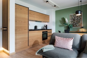 A modern kitchen with wooden cabinets, a black countertop, and a built-in oven. There is a grey sofa with a pink pillow and a green throw blanket. A painting of flowers hangs on the green wall.